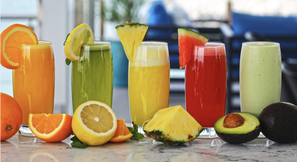 Fives juices you can drink at the start of your juice detox. The juices are orange, green, yellow, red, and green again. They are on a counter surrounded by fruit. 